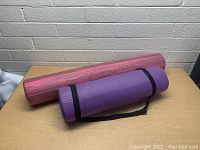Purple yoga mat rolled and secured with a black carrying strap. Red and black marble patterned foam roller placed behind the yoga mat.