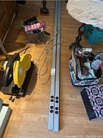 Two long aluminum extension bars lying on wooden floor, each with 4 electrical outlets and attached cords, surrounded by various household items including a saw and tables.