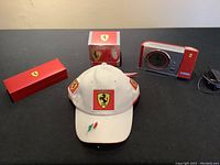 White Ferrari hat with logo patches on front and sides, Italian flag embroidery on brim, placed alongside boxed mug, projection clock, and pen on black surface.