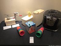 Overview of all items on a black table cloth including apple peeler, air fryer, Tupperware containers, cheese prongs, and popsicle molds