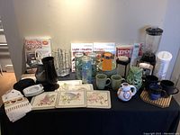 Photo showing entire lot on table including mixer, mugs, ceramic owl holder, water jug, patio cups, cookbooks, and other kitchen items.
