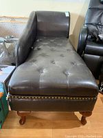 Full view of dark brown faux leather chaise lounge showing tufted seat, curved left armrest, decorative nailhead trim, and wooden legs.