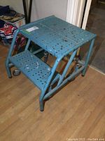 Front angle photo showing two perforated metal steps and top platform, blue paint chipped and worn, positioned on caster wheels.
