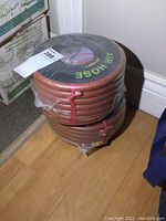 Two orange air hoses rolled and wrapped in plastic with labels showing 3/8 inch x 50 ft dimensions, stacked one on top of the other on a wooden floor.