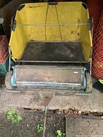 Front view of Parker leaf sweeper showing the sweeping brush and yellow fabric bag, placed indoors on a dirt floor.