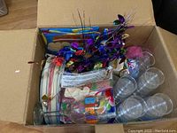 Open cardboard box showing clear plastic glasses, packaged paper plates, balloons, and colorful metallic streamers with red, gold, and purple heart shapes.