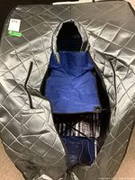 Front view of the portable sauna showing the black quilted exterior fabric with open zipper revealing blue foldable seat inside.