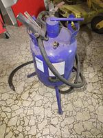 Front-side view of blue 5 gallon sandblaster with hose coiled around it and attached nozzle with trigger handle.