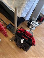 Sledge hammer, pick axe and shovel leaning against the wall with one black and red tool bag in front on wooden floor. The tools show wear.