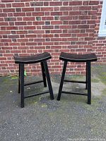 Two black wooden counter stools placed outdoors on pavement with a brick wall background, showing general shape and design