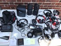 Photo showing multiple wired headphones, earbuds, two black speakers, wired microphones, and cables on table with a brick wall background.