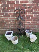 Overall view of the iron gate piece leaning against a brick wall with four decorative planter pots arranged on grass in front.