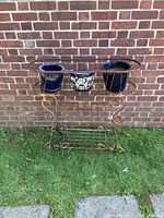 Full front view of rusty vintage wrought iron plant stand with three pots placed on top against a brick wall.