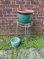 Metal plant stand holding a large green glazed pot with brown rim, smaller green glazed pot with cutouts on the ground in front