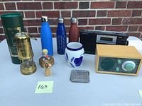 Full lot showing brass Miner's lamp, three water bottles, two old radios, white vase with blue fruit decoration, small figurines, and small silver-tone tray on table with brick wall background.