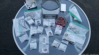 Overview photo of all jewelry pieces spread on a glass table surface, showing multiple bagged chains, bracelets, earrings, and pendants mainly in silver color