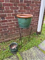Metal plant stand with large green glazed pot on top, small green glazed pot with stones beside it on the grass and stone edge