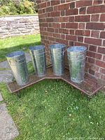 Full view of metal corner plant stand with four galvanized metal flower holders placed on top in an outdoor grassy area near brick wall.