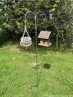 Full view of double shepherd's hook with wooden bird feeder and cream wrought iron hanging planter in outdoor garden setting.