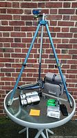 Photo showing the full lot including the tripod, camcorder, camera case, remote, and video tape packs on a round glass table against a brick wall background.