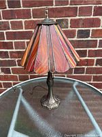 Full view of the stained glass table lamp showing the conical, multi-colored glass shade and detailed silver-tone metal base on a table against a brick wall.