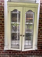 Front view of vintage hanging cabinet with double stained glass doors attached to chains, mounted on brick wall background.
