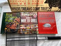 Three BBQ cookbooks, a black metal BBQ grill tool with basket design, and a glimpse of the BBQ sign in the background.