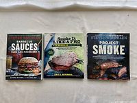 Three barbecue cookbooks laid flat on a surface, showing front covers with images of grilled meats and sandwiches.