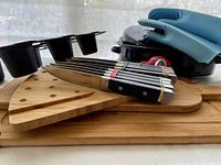 View of knives, cutting boards, and oven mitts stacked, showing knives with black handles and serrated blades, wooden boards, and blue oven mitts.