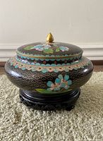 Front side view of small round cloisonné dish with lid on wooden carved stand, showing floral designs and finial on lid.