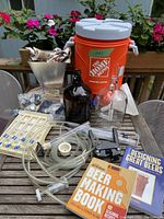 Full view of home beer-making accessory set including orange plastic mash tun cooler, two glass fermenters, plastic tubing, clamps, metal barley grinder, airlocks, and beer-making books