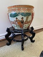 Side view of the ceramic plant pot on black wooden stand, showing painted scene of two women and child.