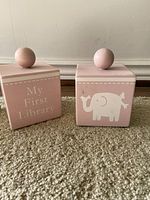 Two pink wooden cube kids' bookends; one with 'My First Library' inscription, the other with an elephant design and round knobs on top, placed on carpet.