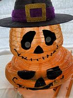 View of lighted stacked jack-o-lantern with facial features, black witch hat with purple band and gold buckle.