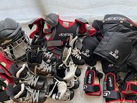 Overall photo showing all items including four pairs of ice skates, two helmets, shoulder pads, gloves, hockey pants, shin pads all arranged on a white surface.
