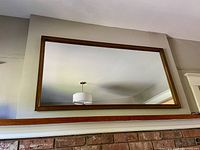 Front view of a large rectangular mirror with a wooden frame hanging above a fireplace mantle, showing the ceiling light reflection.