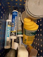 Top view of multiple tubes of caulking including DAP Alex Plus, Dynaflex 230, and others, plus a yellow TILE-Lab grout and tile sealer container inside a perforated crate.