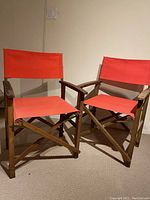 Pair of outdoor folding chairs with wooden frames and red fabric seats and backs against a plain light wall and carpeted floor.
