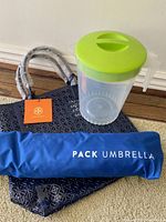 Tory Burch navy blue plastic tote bag, blue umbrella carrying bag labeled 'PACK UMBRELLA', and large clear plastic jug with green lid and handle arranged on floor.