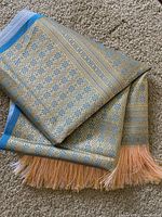 Folded woven blue scarf view showing detailed gold and blue geometric floral patterns and peach tassels.