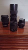 Four camera lenses arranged on wood surface showing Nikon AF NIKKOR 35-80mm, Milda 135mm, FOCAL 80-200mm, and Carenar 28mm lenses in black finish.