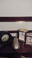 Four small mantle clocks grouped together on a dark wooden surface against a white wall, showing all four clocks clearly including the pewter castle clock, Howard Miller floral clock, wooden antique replica clock, and the black mantle clock.