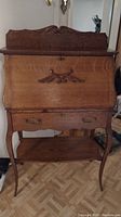 Full frontal image of antique oak slant top desk showing carved top backboard, closed slant top, drawer with brass handles, and curved legs.