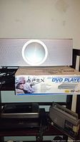Silver speaker unit on top of Apex DVD player box, black device partially visible below, cream colored wall background.