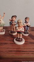 Four figurines on a wooden surface, showing various children in pastel clothing and scarves.
