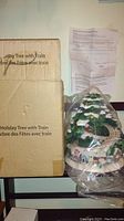 The original box labeled 'Holiday Tree with Train' with instructions visible above the displayed tree figurine wrapped in clear plastic.