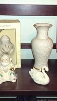 Photo showing a white Lenox bud vase with embossed floral design, a white swan figurine, and a small Santa figurine with green leaves and gold accents
