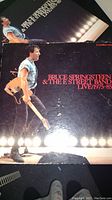 Front cover of Bruce Springsteen & The E Street Band Live 1975-85 3 CD set showing Bruce Springsteen performing live with guitar on stage under spotlights.