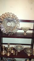 Photo showing large sterling overlay centerpiece bowl on glass with floral design and scalloped edges, two sterling bottom serving dishes, candlesticks, bon bon dish, and Dutch boy and girl salt and pepper shakers on shelf.