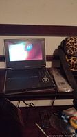Opened Toshiba portable DVD player displaying screen with content. Player is placed on a wooden surface with remote in plastic wrap next to it and a leopard print carrying case.